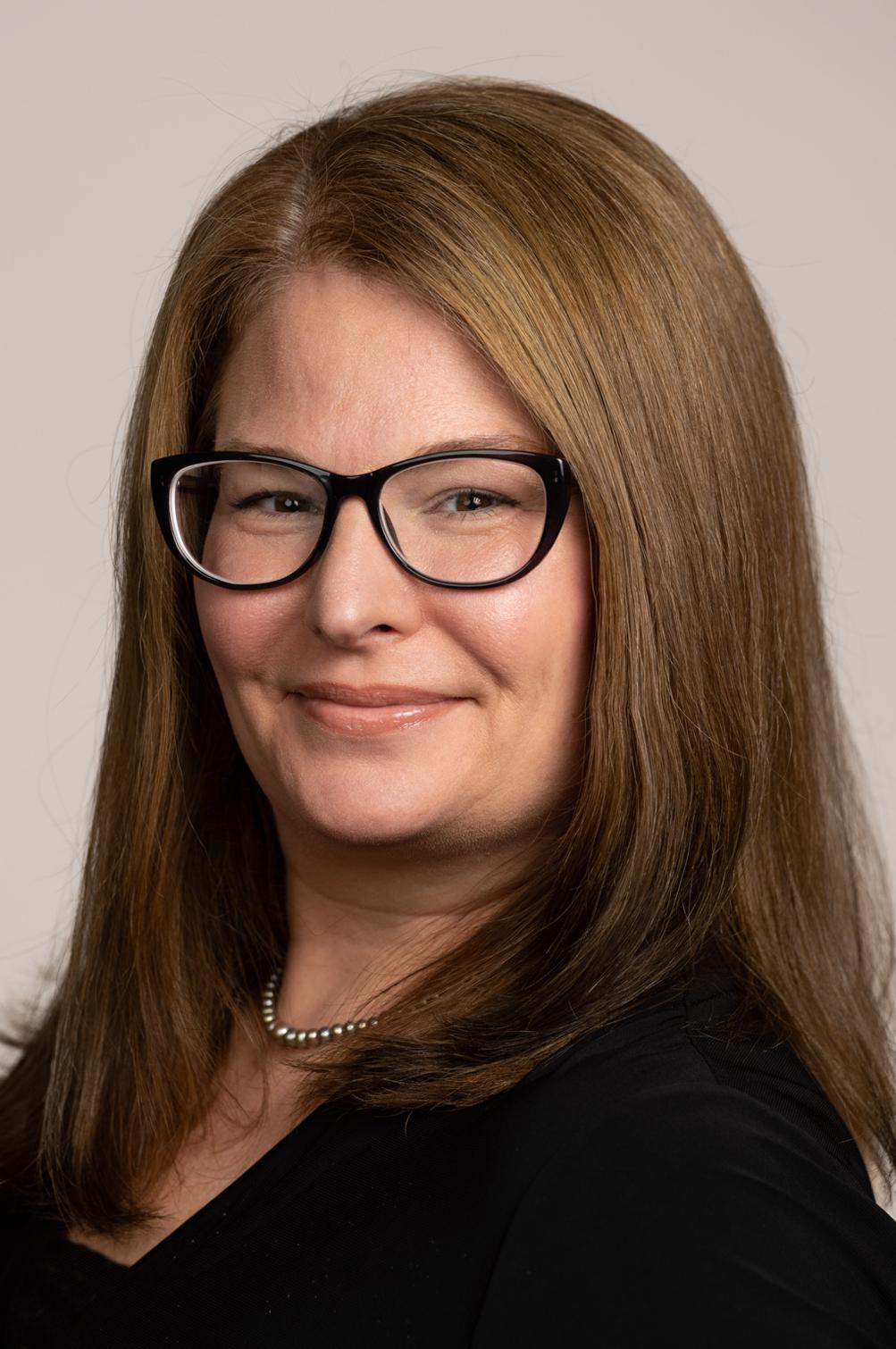 Susan Larsen is a white woman with shoulder-length brown hair and black-rimmed glasses. She is wearing a black top and dark pearl necklace.